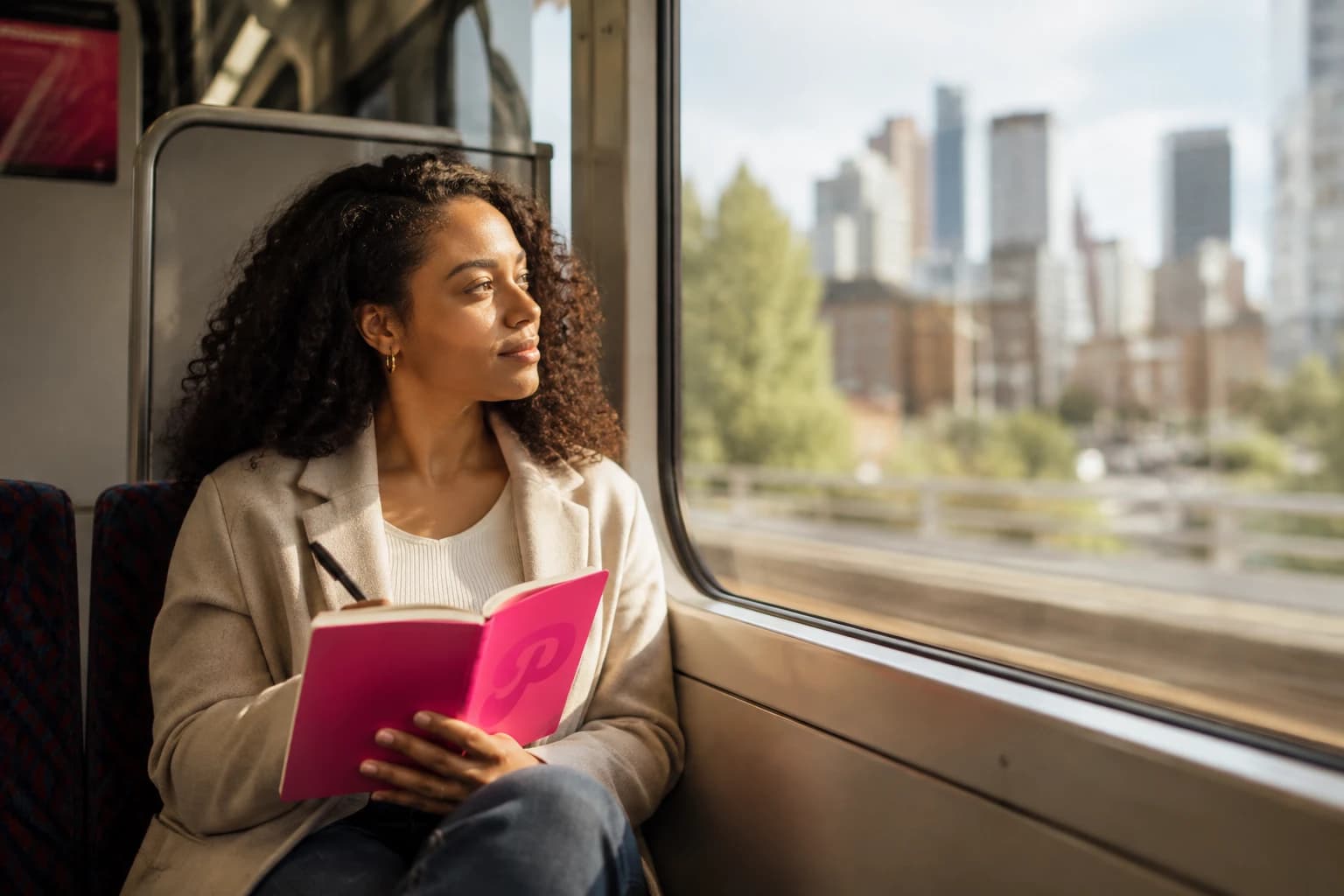 A woman on a train with a pink notebook in her lap, looking out at the city skyline